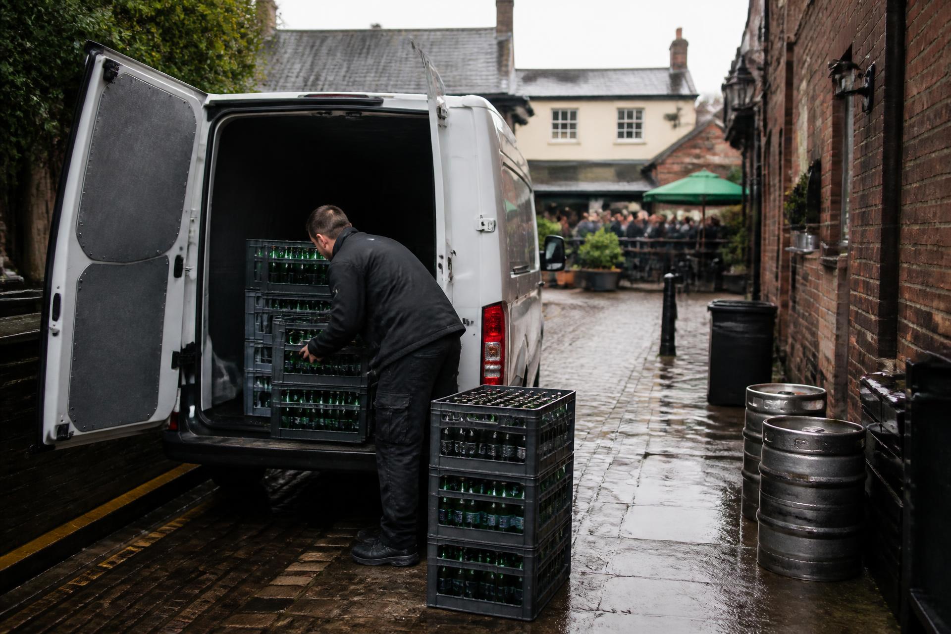 Driver loading crates of green glass bottles into a collection van behind a busy pub